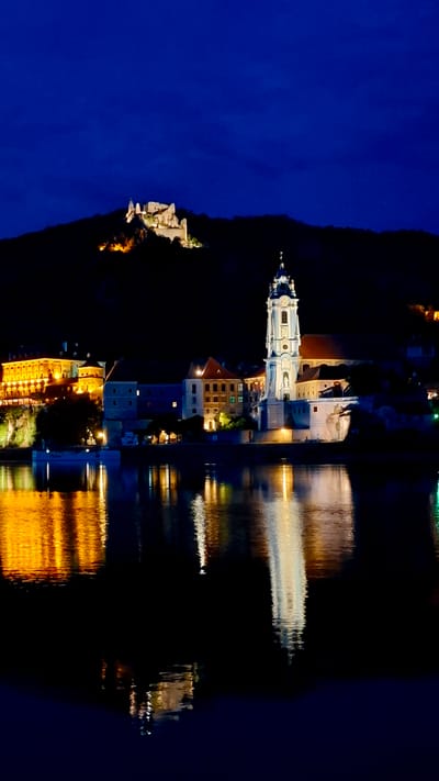 The image is a nighttime view of Rossatz-Arnsdorf, Austria, showcasing the Wachau Valley and the Danube River. In the background, a historic castle ruin is illuminated on a hill. In the midground, a prominent church with a tall, white bell tower stands on the riverbank, flanked by other illuminated buildings. The dark sky is a deep blue, with some faint clouds. The foreground features the dark, reflective surface of the Danube River, mirroring the lights of the town and the church tower. The reflections create streaks of yellow, white, and orange on the water. There are no people visible in the image. The scene depicts a tranquil and picturesque European riverside town at night, with historical landmarks prominently featured.