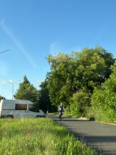 In Kirovo-Chepetsk, Russia, on a sunny day with a clear blue sky, a white van is parked on the left side of a paved road, partially obscured by tall green grass. A man is cycling on the road in the middle ground, wearing a t-shirt and shorts, facing left. Further down the road, a child is also seen cycling, facing left. Dense green trees line the right side of the road, with more trees and foliage visible in the background. Streetlights are present on the left side of the road.