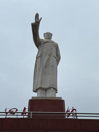 A low-angle, full-shot image captures a tall, light-grey stone statue of a male figure, presumed to be Mao Zedong, against an overcast, light-grey sky. The statue depicts the figure wearing a long, double-breasted overcoat with visible buttons and pockets. His left arm is raised high, palm open, in a gesturing or waving pose. His right arm is bent and positioned behind his back, partially obscured. The figure's head is tilted slightly upward and to the viewer's right. The statue rests on a multi-tiered pedestal. The upper portion of the pedestal is light-grey, matching the statue, while the lower, larger section is composed of reddish-brown rectangular blocks, possibly brick or stone cladding. Below the pedestal, a building or structure with matching reddish-brown facade material is visible. A metal railing, consisting of horizontal bars, extends across the top of this lower structure. Behind the railing, large, stylized red Chinese characters are partially visible, affixed to the top of the reddish-brown structure. The scene is set in Qingyang District, China.