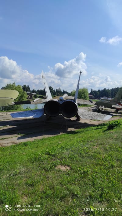 The image shows the rear view of a parked jet aircraft with twin engines. The aircraft is on a concrete pad with grass in the foreground. In the background, there is a body of water, trees, and other buildings. Another aircraft is visible to the right of the main subject. The sky is blue with scattered clouds. The timestamp on the image indicates it was taken on July 15, 2026, at 10:27 AM, and a Redmi Note 9 Pro AI Quad Camera watermark is present.