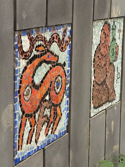 The image displays two mosaic panels embedded in a textured gray concrete wall. The mosaic on the left depicts an orange, stylized animal with large, circular eyes. The animal is positioned diagonally, with its body curving, and appears to have horns or antlers. The background of this panel consists of various shades of blue, white, and brown tiles. The mosaic on the right features a figure in shades of brown and orange, holding a green object that resembles a bottle or flask. This figure is also set against a background of light blue and white tiles. The concrete wall is constructed of large, rectangular blocks. The scene is outdoors, with hints of greenery visible at the bottom right of the frame. The overall impression is of an architectural detail or public art installation. This mosaic is located on the exterior wall of the Borjomi Central Park building in Borjomi, Georgia.