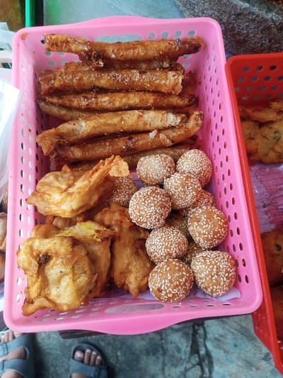 This image displays a vibrant assortment of Indonesian street snacks, characteristic of a food stall or vendor in a location like Cipendawa. The central focus is a pink plastic basket filled with three distinct types of golden-brown fried delicacies.
In the upper section of the pink basket, a stack of elongated, cylindrical items, possibly "pisang molen" (banana wrapped in pastry and fried) or similar sweet spring rolls, gleam with a slightly glossy finish. Below these, on the left, are several pieces of irregularly shaped, battered and fried items, strongly resembling "pisang goreng" (fried banana) or other fritters. The bottom right portion of the pink basket is filled with numerous small, spherical snacks, heavily coated in white sesame seeds, commonly known as "onde-onde" (sesame balls).
Adjacent to the pink basket, partially visible on the right, is a red plastic basket containing more fried items, appearing to be flatter fritters, perhaps "bakwan" (vegetable fritters) or "tempe mendoan" (fried tempeh). The food is brightly lit by natural daylight, indicating an outdoor setting during the daytime. At the bottom of the frame, the feet of at least two individuals wearing sandals are visible on what appears to be a concrete or paved ground, suggesting customers or vendors are standing by. A dark, rough-textured object is visible in the upper right background. The scene effectively captures the everyday culinary offerings of Indonesian street food culture. No text is visible in the image.