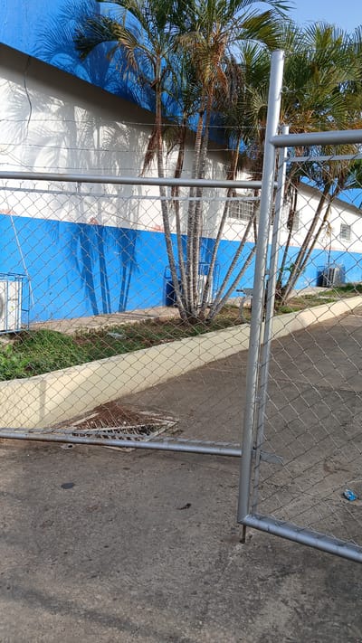 The image shows a partial view of what appears to be a building facade painted with blue and white stripes, potentially indicative of institutional or commercial branding. Several palm trees are visible, with their fronds extending into the frame, suggesting a tropical or subtropical environment. A chain-link fence runs across the foreground, with a metal frame structure partially obstructing the view. The ground surface is concrete, showing signs of wear and debris. A small piece of trash is visible on the concrete. Based on the prompt, the location is Barcelona, Venezuela, although specific landmarks or identifiers are not evident in this limited view to confirm the exact area within the city.