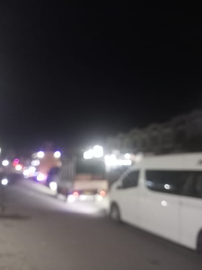 The image is a blurry, low-light photograph taken at night, likely in an outdoor urban setting. The scene appears to be a street with several vehicles present. A prominent white van is visible on the right side of the frame, positioned on a road. Further down the road, a larger truck or bus is partially visible. There are also various lights, some of which appear to be vehicle headlights or streetlights, scattered throughout the scene, contributing to the blurry effect. The background is dark, suggesting it's nighttime, with some indistinct shapes that could be trees or buildings. The overall impression is one of a street scene at night with traffic. The location in Tingkeummanyang, Indonesia, is not visually discernible from the image itself.