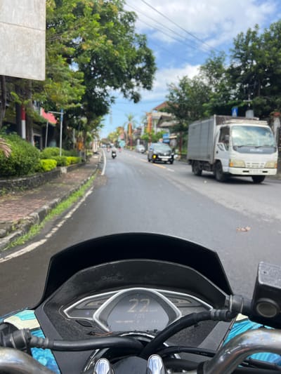 The image captures a point-of-view perspective from a scooter on a street in Klungkung, Indonesia. The road surface is wet, indicating recent rain. Traffic is light, with a white Isuzu truck prominently in the right lane and a dark SUV in the left lane. Several motorbikes are visible further down the road. The sides of the road are lined with lush green vegetation and some buildings. The sky is partly cloudy with blue patches. The scooter's digital dashboard, showing a speed of 22 km/h and other readings, is clearly visible in the foreground. A metallic handlebar with a brake fluid reservoir is also present.