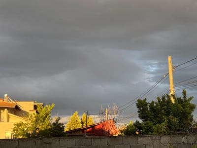 The image shows a residential scene in Manole, Bulgaria under an overcast sky. In the foreground, a concrete block wall spans the width of the image. Behind the wall, various trees and bushes are visible, partially obscuring buildings. To the left, a multi-story beige building with a tiled roof and a solar panel is present. Further to the right, a red-roofed structure is partially visible. An utility pole with numerous wires is positioned to the right of the image, surrounded by lush greenery. The sky is dominated by heavy, dark clouds. No people are visible in the image.