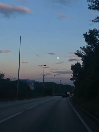 The image captures a tranquil dusk scene along a road, likely in a rural or semi-rural area like Bratica, Montenegro. The sky, transitioning from a soft blue at the top to a muted pink and orange hue near the horizon, is dotted with scattered, elongated clouds. A prominent full or nearly full moon shines brightly in the upper portion of the sky.
Dark, silhouetted trees and dense foliage frame the top and bottom edges of the scene. A paved road stretches into the distance on the right, curving slightly. Along the left side of the road, several streetlights with circular elements are visible, their dark poles extending upwards. In the distance on the road, faint lights from a few vehicles can be seen, with one vehicle closer up displaying visible red taillights. In the mid-ground, a billboard or sign stands by the road, but its content is illegible. Distant hills or mountains are visible on the horizon, blending with the evening light. The overall atmosphere is peaceful and serene. No legible text is visible in the image.