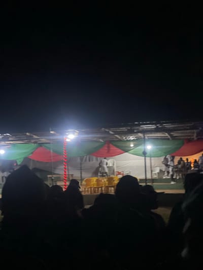 The image depicts an outdoor gathering at night in Orun Ekiti, Nigeria. A tent structure with red and green fabric drapes forms a canopy overhead, illuminated by artificial lights. In the background, a group of people are visible, some standing and others seated on chairs. Yellow plastic chairs are arranged in rows. A vertical string of red lights is visible in the center of the frame. The foreground is filled with the dark silhouettes of people observing the scene, suggesting they are an audience or attendees at an event. The overall atmosphere appears to be festive or ceremonial.