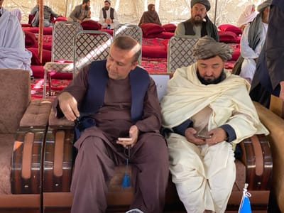 Two men sit side-by-side in ornate armchairs, each focused on a mobile phone. The man on the left wears a brown shalwar kameez and a navy vest, holding prayer beads. The man on the right is wrapped in a cream-colored shawl and wears a turban. A group of people sit on cushions in the background, some wearing turbans and vests. The setting appears to be a tent or covered area with a red floor covering.