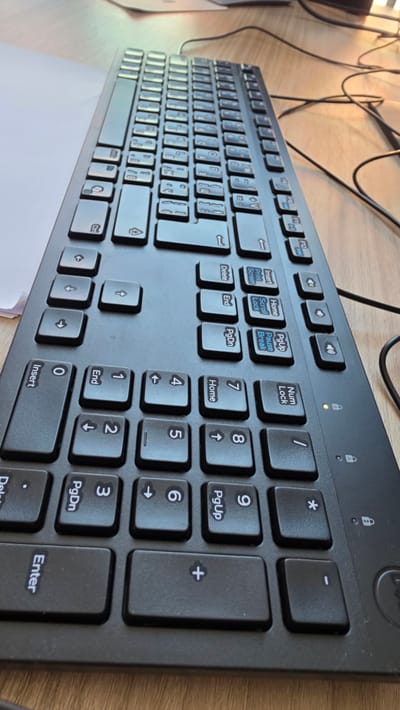 A close-up, overhead shot shows a black computer keyboard on a light brown wooden desk. The keyboard is positioned diagonally from the lower left to the upper right of the frame. The keys are standard QWERTY layout with additional function and navigation keys. Some keys are illuminated with an orange light, indicating activity or status. Several cables are visible trailing from the keyboard to the right side of the frame. The image is taken from a slightly elevated angle, suggesting the camera is held by a person or mounted above the desk. The environment appears to be an office or workspace.