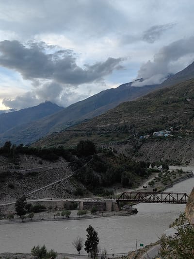 A wide shot captures a river flowing through a mountainous landscape. The river's muddy water runs from left to right across the lower third of the frame. A rusty metal bridge, supported by stone abutments, spans the river. On the far bank, a road winds through sparse vegetation and trees. The opposite bank is a steep, terraced mountainside dotted with trees and a small village with blue-roofed houses. Further back, more mountains rise, their slopes covered in scrub and shadowed by dramatic, grey clouds. The sky is overcast with dramatic cloud formations, suggesting inclement weather or the approach of dusk. The scene appears to be in Gushal, India, a mountainous region known for its natural beauty.