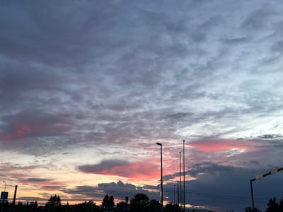 The image depicts a twilight sky over the city of Laagri alevik, Estonia. The sky is filled with dramatic clouds, tinged with shades of gray, purple, and vibrant pink from the setting sun. Several tall streetlights stand silhouetted against the sky, along with power lines. In the foreground, the dark silhouettes of trees and buildings are visible, suggesting a suburban or semi-urban landscape. The overall impression is a moody and atmospheric scene of a sunset in Estonia.