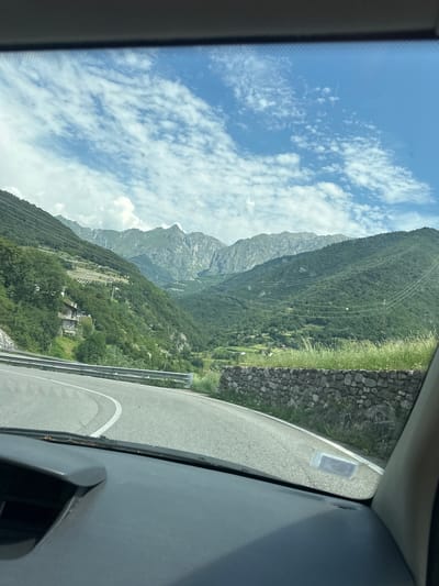 The image shows a view from a car driving on a winding mountain road in Malegno, Italy. The road curves to the left in the foreground, with a metal guardrail along the edge. Beyond the road, a stone wall runs along the side of a grassy embankment. In the background, steep, tree-covered mountains rise towards a bright blue sky with scattered white clouds. Several houses and buildings are visible on the slopes of the mountains, along with power lines strung between poles. The scene appears to be set during the daytime.