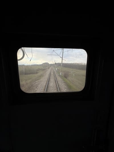 The image displays a view from inside a train, looking forward through a rectangular window with rounded corners. The interior of the train cabin, likely a control car or locomotive, is predominantly dark, framing the brighter outdoor scene.
Through the window, a set of double railway tracks stretches into the distance, converging at the horizon. The tracks are laid on a bed of gravel ballast. On the left side of the tracks, parts of an overhead line (catenary) system are visible, including support structures and electrical wires, indicative of an electrified railway. The surrounding landscape consists of wide, somewhat barren grassy fields with a line of trees or bushes in the distance, suggesting a cold season like late autumn or early spring. The sky is overcast, providing soft, diffused daylight.
No people or vehicles are present in the visible scene. The image portrays a tranquil journey through a rural or semi-rural environment. This scene is located in Urusovo, Russia. There is no visible text in the image.