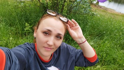 An outdoor, medium-shot, selfie-style photograph features a woman with auburn hair, wearing dark grey long-sleeved shirt with orange trim. She is outdoors in a grassy area with trees and a body of water in the background. She has glasses perched on top of her head and her right hand is raised to adjust them. The woman is looking directly at the camera with a slight smile. A purple balloon is visible in the upper right corner of the image. The overall scene suggests a casual, recreational setting. The specific location is the city of Orekhovo, Russia.