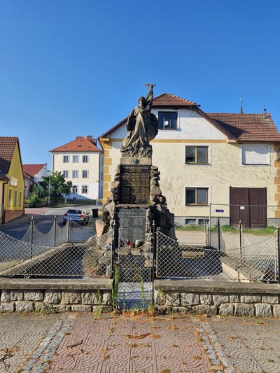 The image shows a war memorial in the town of Střelice, Czechia. The memorial, made of stone and adorned with a statue of a figure holding a bird, is located in front of a light-colored building with a red tiled roof. The memorial is surrounded by a metal fence. In the background, there are other buildings with red tiled roofs, a street with a car parked on it, and a clear blue sky. The foreground shows a cobblestone pavement. The memorial appears to have inscriptions on it, though they are not clearly legible.