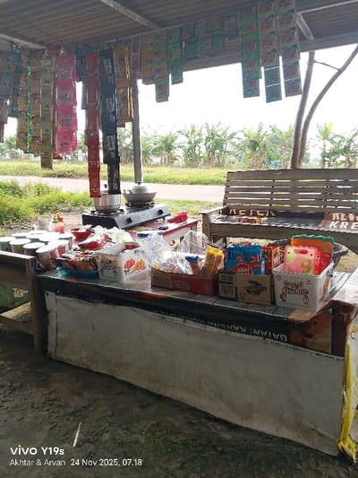 An outdoor food stand displays a variety of snacks and prepared items. Cooking equipment, including pots and a burner, sits on a red table. A bench is nearby. Packaged goods like chips, candies, and possibly fried chicken are arranged on the counter. Hanging from the ceiling are numerous colorful packets of various unknown products. The backdrop features a lush green landscape with banana trees. Text at the bottom indicates "vivo Y19s" and a date.
