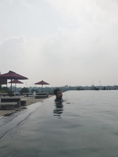 A man is partially submerged in an infinity pool, facing towards the camera with a slight smile. The pool has a concrete edge and a waterfall effect spilling into the foreground. In the background, there are several large red umbrellas, picnic tables with benches, and trees. In the distance, there are two cell towers and a hazy, tree-covered landscape under an overcast sky. The location is identified as Gunung Putri, Indonesia.