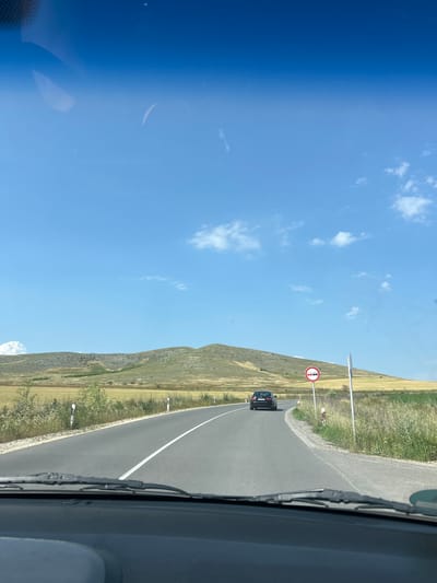 The image depicts a daytime scene from the interior of a vehicle, looking forward through the windshield, with the dashboard and wipers visible in the foreground. The location is Isperihovo, Bulgaria. A two-lane asphalt road, marked with a solid white center line, curves gently to the right. Ahead, a dark-colored sedan, possibly an Audi, travels in the same direction. On the right side of the road, a circular "No Overtaking" regulatory sign, featuring two cars crossed out in red, is mounted on a pole, accompanied by a smaller white delineator post. The landscape beyond the road consists of rolling hills covered in a mix of dry, yellowish-brown grass and green vegetation, extending towards the horizon. Sparse white clouds are scattered across a clear, bright blue sky.