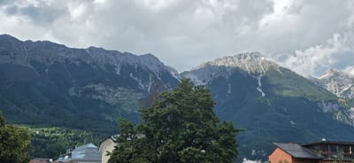 The panoramic image shows a view of the Nordkette mountain range from Innsbruck, Austria. The mountains are covered in dark green forests at their lower slopes, with rocky, lighter-colored peaks and patches of snow visible at higher elevations. The sky is overcast with dramatic grey clouds. In the foreground, a large deciduous tree with dense green foliage is prominent. Below the tree line, several buildings with sloped roofs are visible, including a modern building with a glass facade and balconies, and a smaller orange-colored building. The lighting suggests it is daytime, possibly late afternoon, with some sunlight breaking through the clouds to illuminate parts of the mountainside and the buildings. There are no people visible in the image.