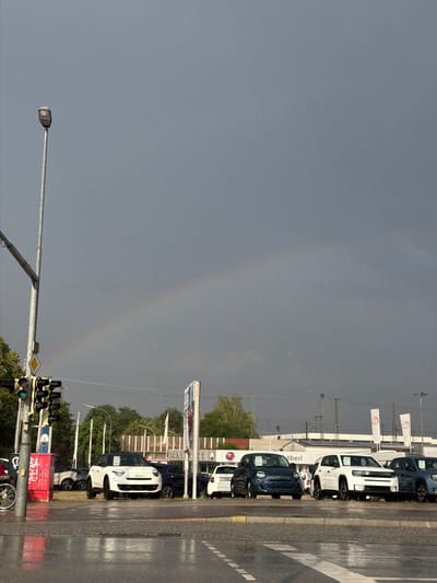 The image captures a car dealership in Ingolstadt, Germany, on a rainy day. The scene is dominated by a row of parked cars, mostly small models like Fiat 500s, in various colors, including white and dark blue. Some cars have price tags or promotional materials visible on their windshields. The ground is wet, indicating recent or ongoing rain, and reflections of the cars and sky can be seen on the asphalt.
In the background, there is a dealership building with signage, including the word "Öberl". Several white flags with a red circular logo are displayed on poles. Above the dealership, power lines and what appear to be train tracks are visible. The sky is overcast and grey, but a faint rainbow arcs across it, suggesting a break in the rain or a recent shower.
In the foreground, a traffic light with green lights illuminated is prominent. Adjacent to it is a red box with white text that is partially obscured but appears to be graffiti or a sticker. A bicycle wheel is also visible at the bottom right. The overall atmosphere is one of a typical urban environment during inclement weather, with a hint of natural beauty from the rainbow. The time of day is not explicitly clear but seems to be daytime due to the ambient light.