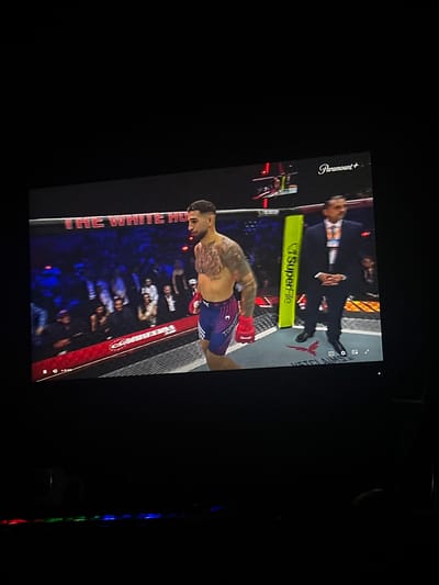 The image shows a screen displaying a fight, likely mixed martial arts, with a fighter in the foreground. The fighter is a male, with tattoos covering his chest and arms, wearing blue and red shorts and red gloves. He is looking to the right of the frame. Behind him, a man in a suit, possibly a referee or official, stands with his hands clasped. The background features a crowd of spectators in the stands. A banner with the text "THE WHITE HO" is visible above the crowd. A logo for "Paramount+" is present in the upper right corner of the screen. The image also shows a dark foreground with what appears to be a keyboard with illuminated keys, suggesting the screen is a monitor. There is no indication of the location being Perímetro Urbano Barranquilla, Colombia.