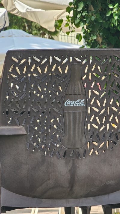 This image shows a close-up view of a dark-colored metal chair with a design of bottle shapes cut out of the backrest. The Coca-Cola logo is prominently embossed in the center of the bottle-shaped cutout, on a larger bottle silhouette. The chair appears to be outdoors, with blurred images of white umbrellas and green foliage in the background. Based on the context provided, this chair is located in Alhama de Aragón, Spain.
