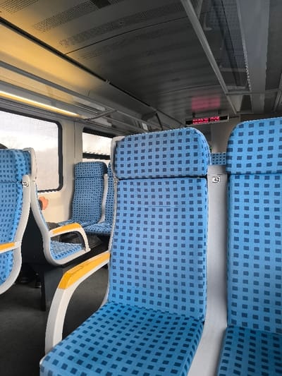 The image displays the interior of a passenger train car located in Kopanitsa, Bulgaria. The scene is dominated by multiple rows of light blue seats featuring a repeating dark blue square pattern. These seats are supported by light grey or white frames, accented with yellow-orange armrests. Some seat frames exhibit numerical labels, such as "42". On the left, large rectangular windows reveal an indistinct, light-colored exterior, suggesting an overcast environment or motion blur. The ceiling is grey and perforated, equipped with linear fluorescent lighting fixtures. An illuminated red LED digital display is mounted on the ceiling, showing "02.03.25 17:05", likely indicating a date and time. A single individual is partially visible in the far-left window seat, with only a portion of their arm and shoulder, clothed in a light-colored sleeve, discernible. The train car appears to be in active use or prepared for operation.