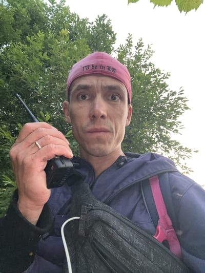 A man in a pink baseball cap with the embroidered text "I'll be in LA" holds a black walkie-talkie in his right hand. He is wearing a dark blue jacket over a black garment and a black fanny pack across his chest. The man's left hand, wearing a ring, is partially visible. He is outdoors, with green foliage and a bright sky in the background. The image appears to be a selfie, taken from a low angle. The location of Badashka, Russia, is not indicated in the image.