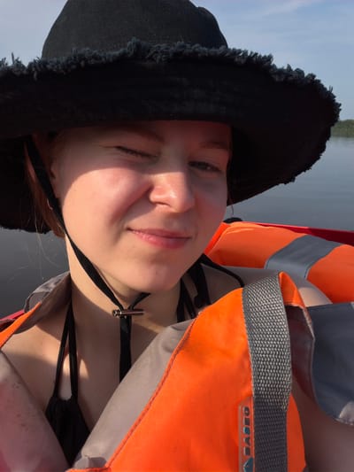 In the city of Kalino, Russia, a young woman with red hair is captured in a close-up, outdoor, chest-up photograph. She is wearing a black, wide-brimmed hat with a frayed edge, a black bikini top, and an orange life vest with gray straps. The woman is positioned on a body of water, with a slightly blurred background of calm water and a distant tree line under a bright sky. Her right eye is closed, and she is looking slightly to the left with a subtle smile. The lighting suggests it is daytime, possibly late afternoon, with sunlight highlighting her face. The image appears to have been taken from a boat or similar watercraft.
