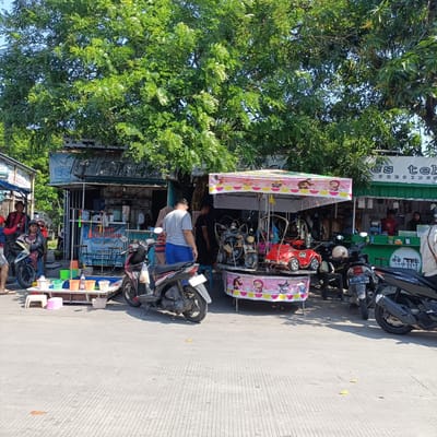 A street scene in Kebondalem, Indonesia, is depicted on a sunny day. In the foreground, a paved area with parallel concrete grooves is visible, with fallen leaves scattered across it. To the left, a person stands near a motorbike, while to the right, several motorbikes are parked. In the mid-ground, a colorful children's carousel ride with toy vehicles like a red car and a yellow motorbike is set up. Adjacent to the carousel is a food stall with green trim and a white sign that reads "Es Teh PRESIDENT". Inside the stall, two individuals appear to be serving customers. Another stall to the left, with a tin roof and a blue tarp, sells drinks. People are milling about near these stalls. A large tree with lush green foliage provides shade over the scene.