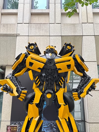 A robot sculpture is positioned outdoors against the backdrop of a tiled building in Unknown, China. The sculpture, constructed from yellow and black materials, resembles the Transformer character Bumblebee. The surrounding environment includes a section of a building facade, a glimpse of tree branches with green leaves, and a sign with Chinese characters and symbols prohibiting climbing. The scene is captured under natural lighting.