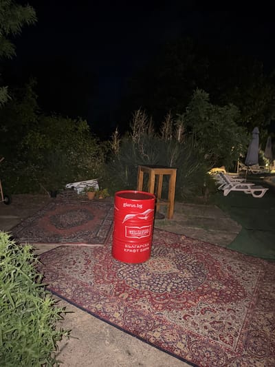 In Varvara, Bulgaria, at night, a red barrel with the text "glarus.bg CRAFT BREWING CO." is positioned on a patterned rug. Adjacent to it, a wooden stool stands. In the background, a swimming pool area is visible with lounge chairs and umbrellas. The scene is set outdoors amidst vegetation and is illuminated by artificial light.