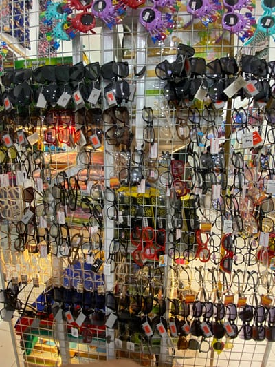 This media file showcases a vibrant display of eyewear, likely sunglasses and eyeglasses, organized on metal racks. The items are varied in style and color, with many pairs featuring dark lenses. Some glasses are attached to colorful, novelty party glasses, such as purple ones with designs resembling flowers or a character's head, and red ones with decorative elements. There are also individual pairs of glasses in various frames, including black, clear, and patterned. Several price tags and labels with text such as "ROLLIES," "POLAR BUND," "ORIGINAL," and "KIDS" are visible, along with numerical price indications like "30k" and "25k." The background suggests an indoor market or store setting, with hints of colorful fabrics or displays behind the eyewear. The lighting appears to be artificial, casting a warm glow on the merchandise, suggesting it might be daytime or evening within a well-lit retail space. The overall impression is one of a bustling point of sale, with a wide assortment of affordable eyewear available to customers. The location context provided is Lamteumen Barat, Indonesia.