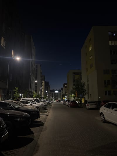 The photograph depicts an urban street scene at night in Sokilnyky, Ukraine. The street is lined with parked cars on the right side, and buildings rise on both sides. The sky is dark, suggesting it is late evening or night. Streetlights illuminate the scene, casting shadows along the road and highlighting the facades of the buildings. The road is paved with bricks. There is no apparent activity, interaction, or people.