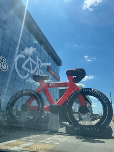 This is a photograph taken outdoors in Yablonovskiy, Russia, on a sunny day with scattered clouds. The primary subject is a large, inflatable red and black bicycle sculpture positioned in front of a modern glass-fronted building. The building has a large bicycle logo displayed on its facade, reflecting the sky and clouds. Red signage on the building reads "ВЕЛОСИПЕДЫ" (Bicycles) and "Gukecenter" with additional text in Russian. The inflatable bicycle has "В НАЛИЧИИ" (In Stock) and "BIKE-CENTRE.RU" printed on its large black hoop, resembling a wheel. Another section of the hoop has text that appears to be a phone number: "8 800 500 03 20". The overall scene suggests a commercial setting, likely a bicycle store or related business, emphasizing availability and a strong brand presence. The bright sunlight casts shadows, indicating it is likely daytime. The image appears to be taken from a slightly low angle, looking up towards the inflatable sculpture and the building.