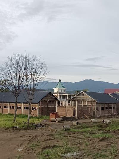This is a medium-shot, eye-level, outdoor photograph of a construction site on a cloudy day in Beurawang, Indonesia. In the foreground, there is a muddy, uneven ground with patches of green grass and several large, cylindrical concrete pipes lying on their sides. Behind this, there are buildings under construction. One building is mostly complete with a dark, sloped roof and brick walls with arched windows. Another section of the building is still in its framework stage, with exposed wooden beams and brickwork. To the left, two bare, deciduous trees stand in front of a building with a dark roof and a row of windows. In the background, a larger, dome-shaped building with a green roof and white facade is visible, possibly a religious building or a significant institutional structure. Further back, a range of blue, mountainous terrain stretches across the horizon under a heavily clouded sky. The overall scene depicts an active construction area, likely a new development or expansion project in a semi-rural or suburban setting, given the presence of mountains and natural vegetation alongside the built environment.