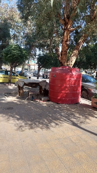 The image depicts an outdoor urban scene within Al-Qaboun Municipality, Syria, under bright daylight. In the foreground, a prominent red, ribbed, cylindrical plastic water tank stands upright on the right, positioned adjacent to a tree trunk. A metallic faucet or valve is visible on the tank's upper side. To the left of the tank, a crude, open-sided wooden structure resembling a stall or table is present, covered by an off-white or light-colored fabric or tarp. Beneath this structure, on the dirt ground, a small blue object, possibly a pump, is visible along with associated hoses or cables.

The immediate ground surface is composed of light-colored, patterned paving stones, transitioning to dirt patches and disturbed earth around the wooden structure and the base of the water tank. Shadows from overhead trees are cast prominently across the paved area.

In the midground, a paved street or road extends into the distance. A yellow sedan is partially visible on the left, parked behind the wooden structure. To the right of the water tank, a grey sedan is parked, partially obscured by foliage and the tank itself. A person, largely obscured, is standing near the grey sedan on the far right. Multiple other vehicles, including cars and possibly a truck, are discernible further down the street. The scene is heavily populated by various trees with green foliage and visible brown trunks, contributing to the overhead canopy and shadows. Buildings with light-colored facades are visible in the background, interspersed with more trees. The sky, visible through the tree canopy, is clear blue.