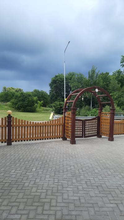 The image captures a frontal view of an outdoor scene with a paved walkway in the foreground. A low wooden fence with rounded tops extends across the left and center of the frame, topped with a rounded finial on a post at the far left. To the right of the fence, a wooden archway with a gate stands, featuring two wooden panels with a lattice pattern. The archway is made of dark red wood, with two vertical posts supporting a curved beam. Hanging from the arch are several small, bell-like objects.
Behind the fence and archway, a grassy area is visible, sloping upwards. In the background, a dense line of green trees forms a natural backdrop. Above the trees, the sky is overcast with dark, heavy clouds, suggesting impending rain. A tall, white lamppost stands to the right of the archway, partially obscured by trees. A metal railing is also visible in the middle ground, running behind the fence. The ground is paved with grey interlocking bricks laid in a herringbone pattern. There are no people visible in the image. The scene appears to be in a park or a landscaped outdoor area.