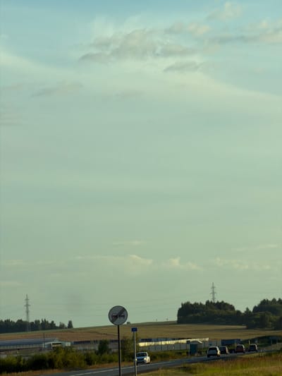 The image shows a rural scene with a road, cars, and a traffic sign. In the foreground, a road curves to the right, with several cars driving on it. A circular traffic sign with a red border and two red cars inside, indicating "no overtaking," is positioned on a pole to the right of the road. In the background, there are fields, trees, and a power line tower. The sky is light blue with some clouds. The overall impression is one of a sunny day in a rural setting.