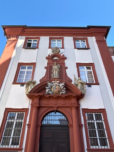 The image shows a building in Heidelberg, Germany. The building has a white facade with red trim around the windows and edges. There are six visible rectangular windows with white frames and multiple panes. Above the main entrance is an elaborate architectural embellishment featuring sculptures, a coat of arms, and a statue. The entrance itself has a large, arched window above a dark-colored door. The sky is visible in the upper left corner and is a clear, light blue.