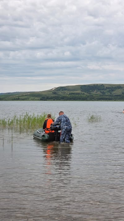 Two individuals are in a dark green inflatable boat on a body of water. One person, wearing an orange life vest over dark clothing, is seated in the boat facing away from the camera. The second person, wearing a camouflage uniform, is standing in the water behind the boat and appears to be adjusting or operating a small outboard motor attached to the stern. The boat is partially submerged, indicating it may be near the shore or in shallow water. Tall green reeds are visible along the left edge of the water, extending towards the boat. The background features a wide expanse of water, with a gently sloping, treed hillside in the distance under a cloudy sky. A road is visible winding its way up the hillside. The overall scene suggests an outdoor activity on a lake or large river, possibly related to recreation or work.