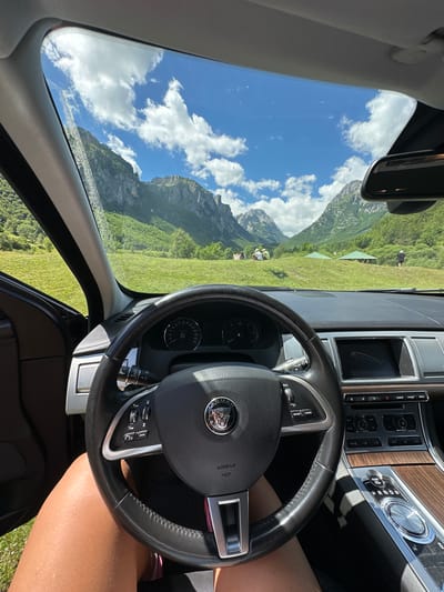 The image is taken from inside a car, looking out through the windshield at a mountainous landscape in Vusanje, Montenegro. The foreground shows the car's interior, including the steering wheel with a Jaguar logo, the dashboard with a digital screen displaying a Jaguar emblem, and the gear shift. A person's legs, wearing shorts, are visible on the left side of the frame. Outside, a lush green meadow slopes up towards a range of majestic, rocky mountains. The sky is a vibrant blue with fluffy white clouds. In the distance, several people are seen relaxing on the grassy hillside, with some sitting and others standing near two green gazebos. The scene depicts a serene outdoor setting enjoyed from the vantage point of a car.
