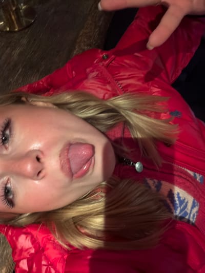 An indoor, close-up photograph depicts a fair-skinned individual with blonde hair, positioned diagonally in the frame with their head tilted. The subject is sticking their tongue out of their mouth. Their eyes are open, with visible dark eyelashes. They are wearing a shiny red quilted jacket, possibly a puffer, with a blue embroidered logo on the left chest area. Underneath, a red garment with a white and blue patterned design is visible, secured with a zipper featuring a silver-colored pull. In the upper right quadrant, the tips of two fingers, possibly forming a "peace" sign, are visible with manicured nails, extending into the frame. The background is dark, revealing a portion of a dark wooden surface in the upper left corner, where a metallic, brass-toned object is partially visible. The lighting is dim. The specific location within the city of La Clusaz, France, cannot be determined from the visual evidence presented in the image.