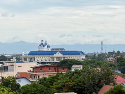 This is a photograph of a sprawling cityscape, dominated by a large, ornate building with a bright blue roof and two prominent blue domes. The building features white columns and detailed architectural accents. In the foreground, several residential buildings are visible, varying in color and style, with red-tiled roofs being a common feature. Lush green trees and foliage are interspersed throughout the scene, suggesting a tropical environment. In the background, mountains are visible on the horizon, partially obscured by a hazy sky. Several telecommunications towers are also scattered across the landscape, indicating the presence of modern infrastructure. The overall impression is of a city that blends traditional and modern elements, set against a natural backdrop. The building with the blue domes is likely a significant landmark in Vientiane Capital, Laos, possibly a government building, a hotel, or a cultural center. The residential buildings suggest a typical urban or suburban neighborhood. The presence of the telecommunication towers points to ongoing development and connectivity within the city. The clear, though slightly overcast, sky suggests a daytime scene, likely taken from an elevated vantage point to capture the breadth of the cityscape.