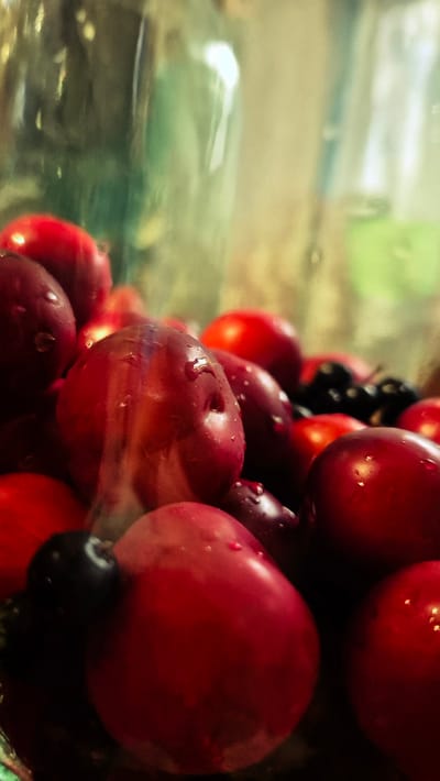 A close-up, shallow depth-of-field photograph captures a collection of ripe, red plums with water droplets scattered across their surfaces, contained within a clear glass jar. A few dark berries, possibly blueberries or blackberries, are interspersed among the plums. The lighting suggests an indoor setting, with reflections visible on the jar and the fruit. The background is blurred, showing indistinct green and beige tones. The image does not contain any recognizable landmarks or elements that would allow for the localization of the scene within the city of Troițcoe, Moldova.