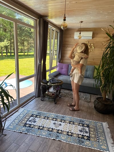 This image captures an indoor-outdoor living space in Копнино, Russia, characterized by extensive natural light and a rustic aesthetic. The scene features one adult female holding a young child, standing on a wooden floor in front of a grey sofa.
The adult female, with long light hair, wears a light-colored t-shirt, matching shorts, and brown slides. She holds a young child, likely a toddler, dressed in a light-colored bucket hat, t-shirt, and striped shorts. The adult's left hand is near the child's hat. The child is facing and embracing the adult.
The interior space features light-colored wood paneling on the walls and ceiling. Two pendant lights with large, exposed Edison-style bulbs hang from the ceiling. A white wall-mounted air conditioning unit is positioned on the wood-paneled wall. Below the air conditioner, a metallic wall decoration resembling golden leaves or butterflies is affixed.
Along the left and back walls, large floor-to-ceiling glass windows and doors offer a clear view of a lush green lawn and numerous trees under bright sunlight. Grey curtains with tie-backs are drawn to the side of the windows. Outside, a wooden deck is visible, with a light blue circular object (possibly a pool cover or small pool) placed on it.
To the left of the sofa, an ornate, two-tiered metal and glass side table is present. On its top tier, a black portable speaker and a folded dark garment are placed. The bottom tier holds a small wooden chess set box and a red box. A small grey faux-fur rug is positioned beneath the side table and partially under the sofa.
A patterned rug, primarily white with blue, grey, and yellow geometric and stylized tree designs, is centrally placed on the wooden floor. To the right of the sofa, a large potted plant with abundant green foliage stands in a dark, textured pot. Part of another plant is visible in a similar pot in the bottom left foreground.