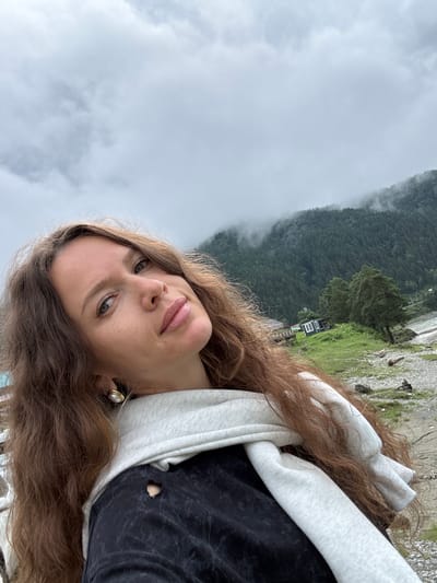 An outdoor, eye-level, medium shot captures a woman in a selfie with a slightly overcast sky and a mist-covered mountainous landscape in the background. The woman is positioned in the lower left of the frame, facing slightly away from the camera with her head tilted back. She has long, wavy brown hair and is wearing a light gray scarf over a black sweatshirt with a torn detail on the left shoulder. She has on a large, pearl-like earring in her left ear. The background depicts a densely wooded mountainside partially obscured by mist, with a body of water to the right and a small, dark-colored building with white window frames situated on a grassy area between the trees and the water. The foreground consists of a rocky and gravelly shoreline. The location is identified as Elekmonar, Russia.