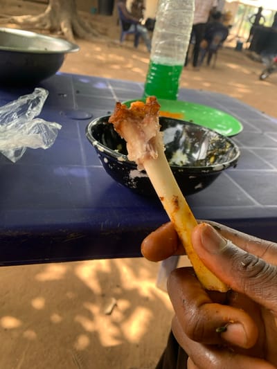 A close-up image depicts a dark-skinned hand holding a partially consumed, yellowish-white bone, likely from a chicken drumstick, with residual crispy meat and spice residue on the fingers. The hand is positioned over a blue plastic table with a textured surface. On the table, a black bowl with white speckles contains dark liquid and food remnants. A crumpled clear plastic bag is also present. In the mid-ground on the table, a clear plastic water bottle with green liquid at its base stands next to a bright green plastic plate. The background reveals an outdoor setting with dry, reddish-brown earth illuminated by dappled sunlight and shadows from overhead trees. Several blurred figures are visible, seated on blue plastic chairs around other tables, suggesting a communal dining area. The scene is located in Assai Sabongida, Nigeria.
