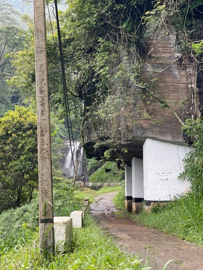 The image depicts a natural scene in Ramboda, Sri Lanka. A waterfall cascades down a verdant, mountainous landscape. A man-made structure consisting of a white, rectangular building with pillars built into the side of a large rocky face, and a path winding through foliage, leads to a view of the falls. Green vegetation surrounds the path and the building. A utility pole with wires is visible in the foreground. The path appears muddy. The building has some writing in an unfamiliar script on one side. The sky is overcast and hazy.